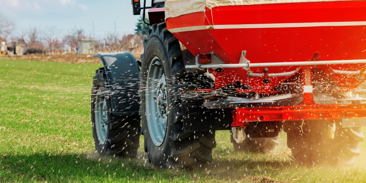 Unrecognizable farmer in agricultural tractor is fertilizing wheat crop field with NPK fertilizer nutrients